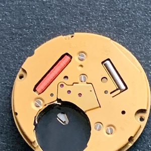 May include: Close-up of a watch movement with a gold-colored base. It features a red rectangular component, a silver-colored component, and several small screws. The watch movement has a circular shape with a dark hole in the center.