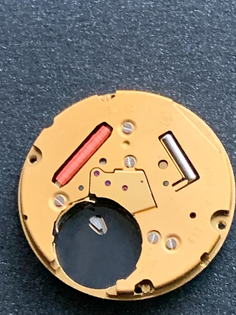 May include: Close-up of a watch movement with a gold-colored base. It features a red rectangular component, a silver-colored component, and several small screws. The watch movement has a circular shape with a dark hole in the center.