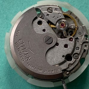 May include: Close-up of a Citizen Watch Co. watch movement. The silver-toned mechanism features intricate gears, a balance wheel, and the text "TWENTY-ONE 21 JEWELS." The watch movement is set against a light green background.