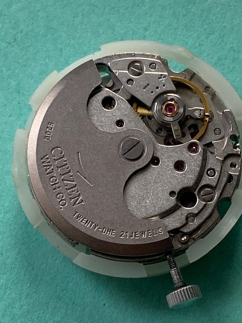 May include: Close-up of a Citizen Watch Co. watch movement. The silver-toned mechanism features intricate gears, a balance wheel, and the text "TWENTY-ONE 21 JEWELS." The watch movement is set against a light green background.