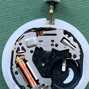 K&ouml;nnte beinhalten: Nahaufnahme eines Uhrwerks mit silberner Krone und wei&szlig;em Kunststoffgeh&auml;use. Die inneren Komponenten umfassen eine Kupferspule, eine schwarze Batterie und verschiedene Metallplatten. Der Text "10 JEWELS UNADJUSTED" ist sichtbar.