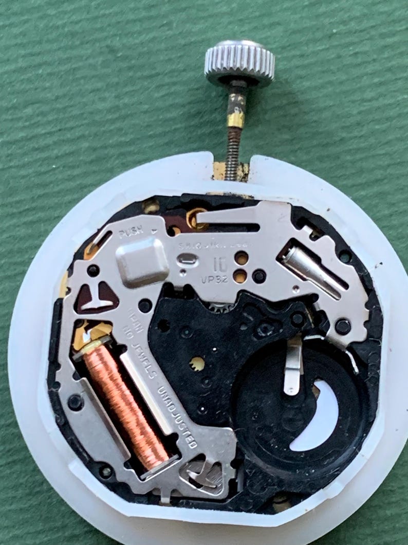 K&ouml;nnte beinhalten: Nahaufnahme eines Uhrwerks mit silberner Krone und wei&szlig;em Kunststoffgeh&auml;use. Die inneren Komponenten umfassen eine Kupferspule, eine schwarze Batterie und verschiedene Metallplatten. Der Text "10 JEWELS UNADJUSTED" ist sichtbar.