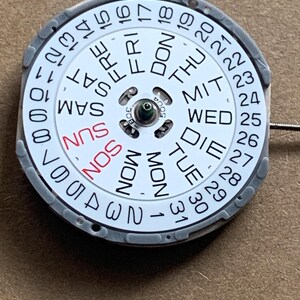 May include: A watch calendar wheel with a white face, featuring black numbers and day abbreviations. The days of the week are in English, with "SUN" in red. The wheel is encased in a gray frame.