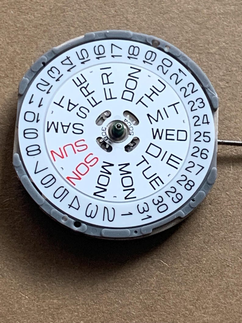 May include: A watch calendar wheel with a white face, featuring black numbers and day abbreviations. The days of the week are in English, with "SUN" in red. The wheel is encased in a gray frame.