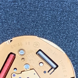 May include: Close-up of a watch movement with gold-colored components. The text "SWISS V8" and "F05 111" are visible. The movement also features a red coil and the text "THREE 3 JEWEL".
