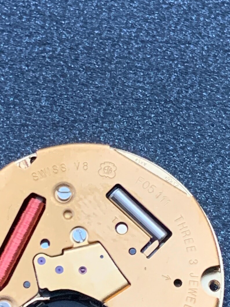 May include: Close-up of a watch movement with gold-colored components. The text "SWISS V8" and "F05 111" are visible. The movement also features a red coil and the text "THREE 3 JEWEL".