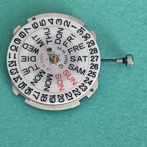 May include: A watch calendar wheel with a white face and black numerals and text. The days of the week are printed in black, with "SUN" in red. The wheel has a silver metal frame and a small screw attached.