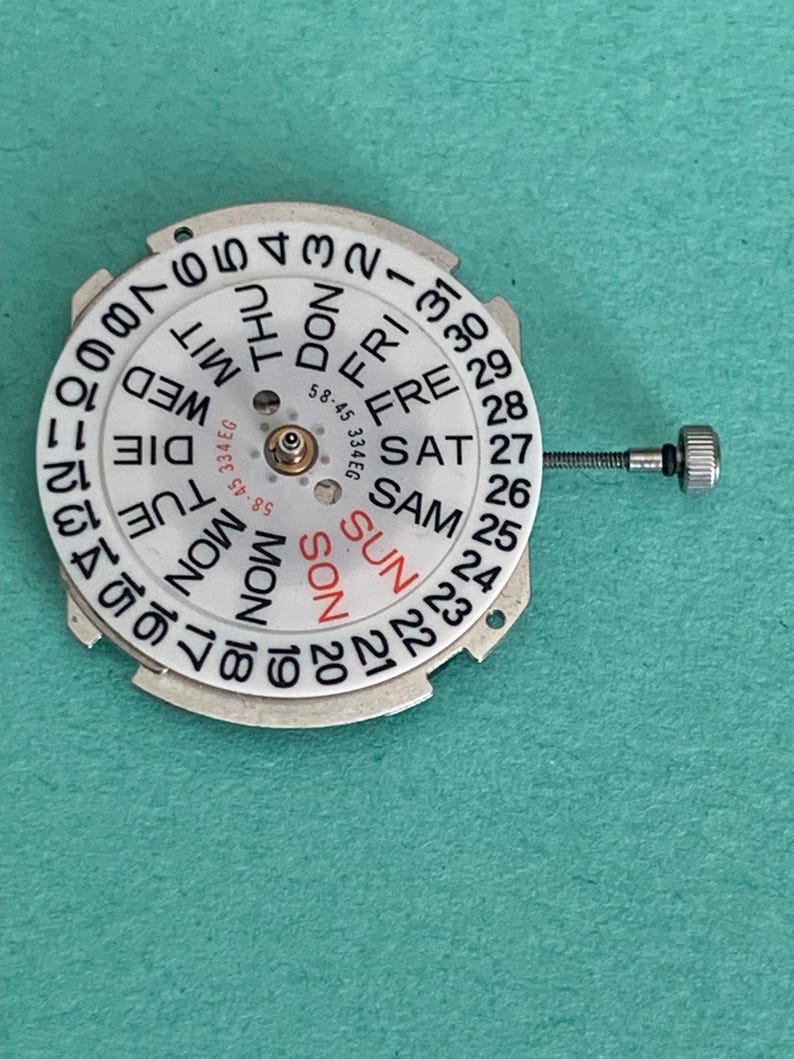 May include: A watch calendar wheel with a white face and black numerals and text. The days of the week are printed in black, with "SUN" in red. The wheel has a silver metal frame and a small screw attached.