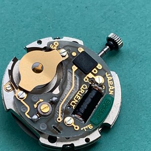May include: Close-up of a watch movement with gold and silver components. The watch mechanism has a black coil, a small black square, and the word "ORIENT" printed on it. The text "JAPAN" and "52550" are also visible.