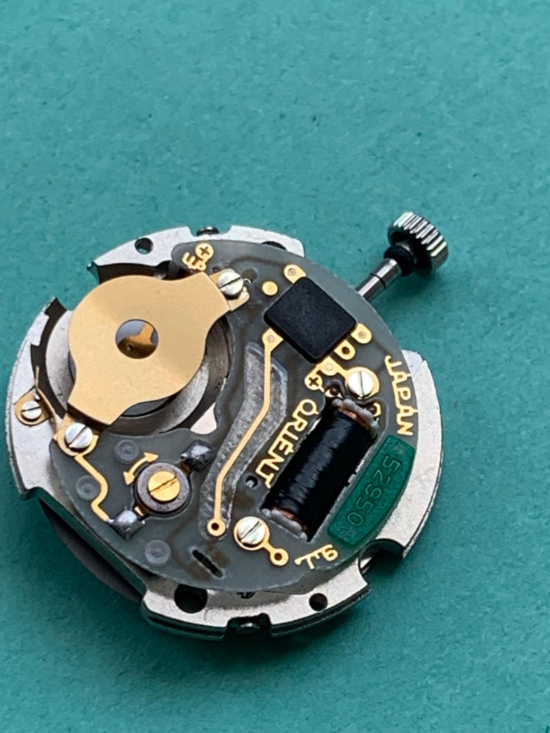 May include: Close-up of a watch movement with gold and silver components. The watch mechanism has a black coil, a small black square, and the word "ORIENT" printed on it. The text "JAPAN" and "52550" are also visible.