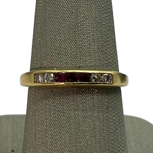 May include: A gold band ring featuring alternating white and red gemstones. The ring has a simple, elegant design with the gemstones set in a row across the top. The ring is displayed on a gray ring holder.