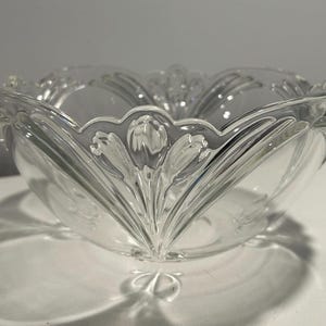 May include: A clear glass bowl with a scalloped rim and a floral design. The bowl's sides feature vertical, petal-like patterns that converge at the base. The bowl is transparent, allowing light to pass through, and casts a shadow.
