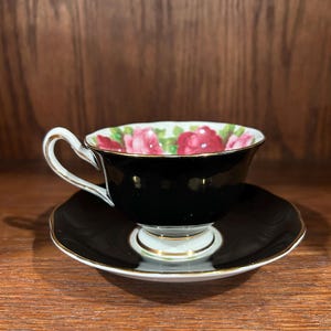 May include: Black teacup and saucer set with a floral rose pattern inside the cup. The cup and saucer have a gold trim. The teacup is on a white base.