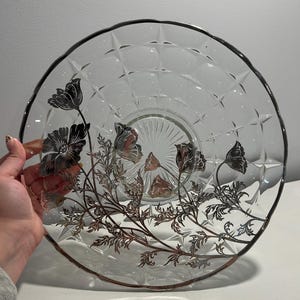 May include: Clear glass serving plate with a silver floral design. The plate has a diamond pattern and a silver rim. The floral design features poppies and stems. The plate is round and appears to be vintage.