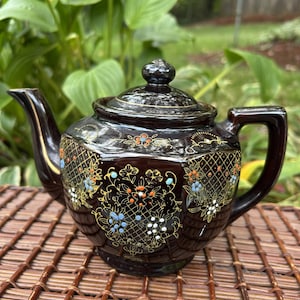May include: A dark brown ceramic teapot with a hexagonal body and a matching lid. The surface features a gold lattice pattern overlaid with hand-painted floral motifs in orange, blue, and white, accented by delicate gold line work and detailing.