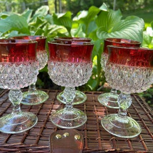May include: Six vintage wine glasses with ruby red rims and diamond-cut patterned bowls. The glasses are arranged on a woven wicker tray, with a leather tag in the foreground. The background features lush green foliage.