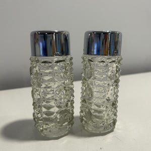 May include: A pair of vintage salt and pepper shakers. The clear glass shakers have a textured, diamond-like pattern and are topped with shiny, silver-colored metal lids. The shakers are cylindrical in shape and sit on a white surface.