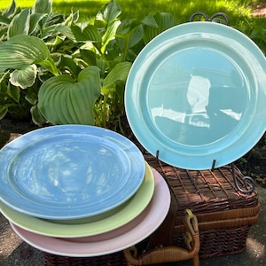 May include: A set of four round ceramic dinner plates in pastel colors. One plate is light blue, one is light green, one is light pink, and one is a larger light turquoise plate displayed upright on a black metal stand.