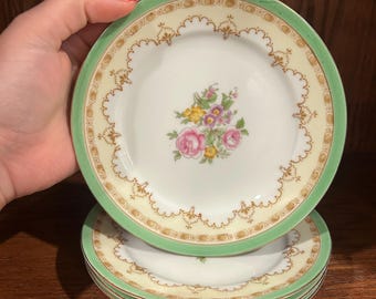 May include: A stack of vintage porcelain plates. Each plate features a floral center design with pink, yellow, and purple flowers. The plates have a light green border and a gold trim. The plates are stacked on top of each other.