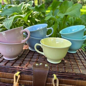 May include: A collection of five ceramic tea cups in pastel colors including light pink, soft blue, and pale yellow. Each cup features a rounded bowl shape, a small pedestal base, and a curved handle, arranged in a group on a woven surface.