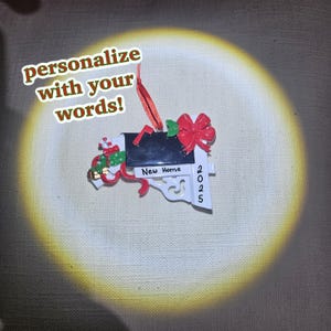 Puede incluir: Adorno navideño de buzón blanco con cinta roja, lazo y detalles verdes. El adorno tiene una puerta de buzón negra y el texto "New Home 2025". También está presente el texto "personalize with your words!"