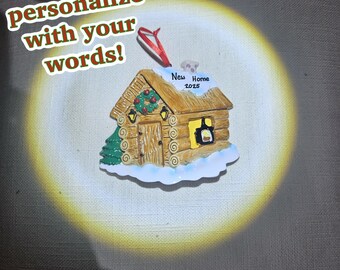 May include: A cabin-shaped Christmas ornament with the text "New Home 2025." The ornament is brown with a green Christmas tree and a wreath. The text "Personalize with your words!" is also visible.
