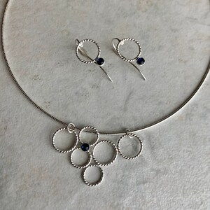May include: A silver necklace with a pendant made of multiple intertwined circles, each with a small blue gemstone. Matching earrings with a single circle and blue gemstone.