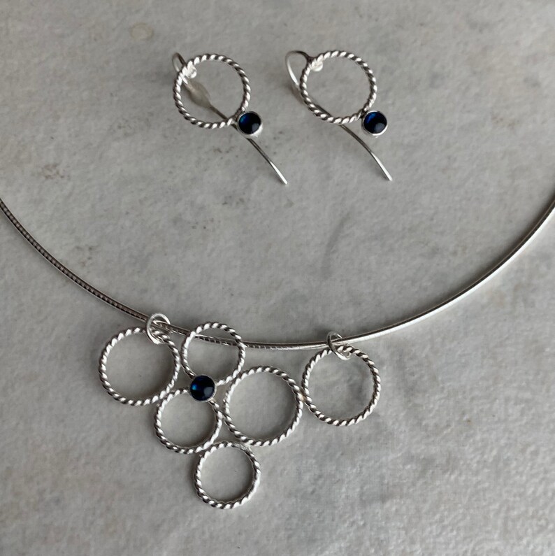 May include: A silver necklace with a pendant made of multiple intertwined circles, each with a small blue gemstone. Matching earrings with a single circle and blue gemstone.