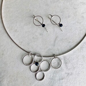 May include: A silver necklace with a pendant made of multiple interconnected circles, each with a small black gemstone. Matching earrings with a single circle and black gemstone dangle from silver posts.