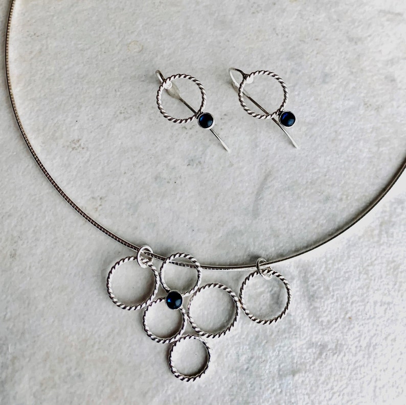 May include: A silver necklace with a pendant made of multiple interconnected circles, each with a small black gemstone. Matching earrings with a single circle and black gemstone dangle from silver posts.