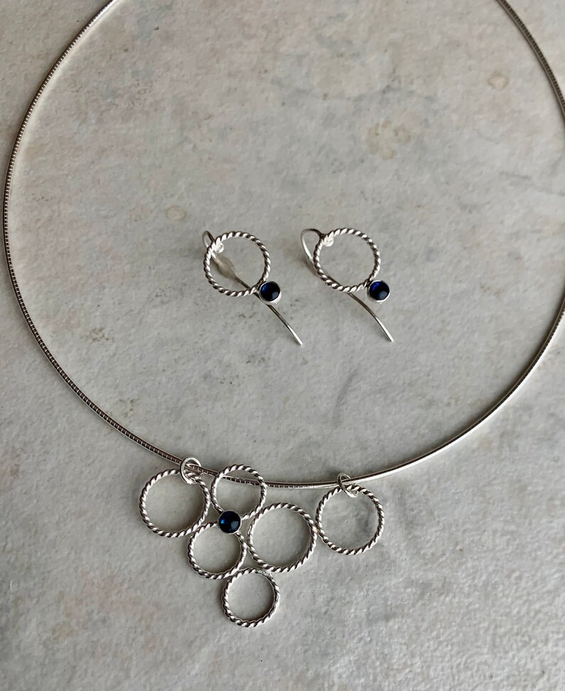 May include: A silver necklace with a pendant made of multiple intertwined circles, each with a small blue gemstone. Matching earrings with a single circle and blue gemstone.