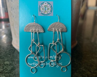 May include: Silver earrings with a geometric design. The earrings feature a half-circle top with a textured surface and dangle with multiple geometric shapes. The earrings are displayed on a turquoise card with the text "Turtle Jewelry Designs".