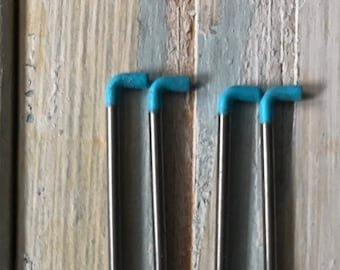 Może przedstawiać: Cztery srebrne druty do robótek ręcznych z niebieskimi plastikowymi końcówkami. Druty są ułożone pionowo na drewnianej powierzchni.