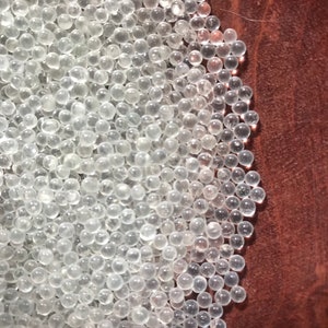 May include: A close-up image of a pile of clear silica gel beads. The beads are small and round, and they are arranged in a random pattern. The beads are on a brown surface.