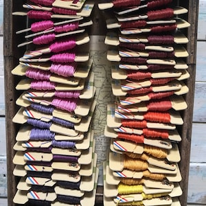 May include: A wooden box filled with spools of colourful embroidery floss. The floss is organised by colour, with shades of pink, purple, red, orange, and yellow.