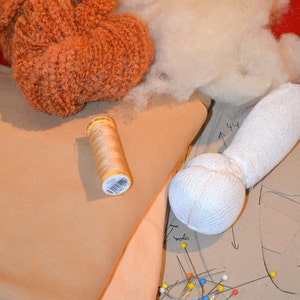 May include: A close-up of a craft project with a spool of thread, a white fabric ball, and a piece of beige fabric. There are also colorful pins and a pattern on the fabric.