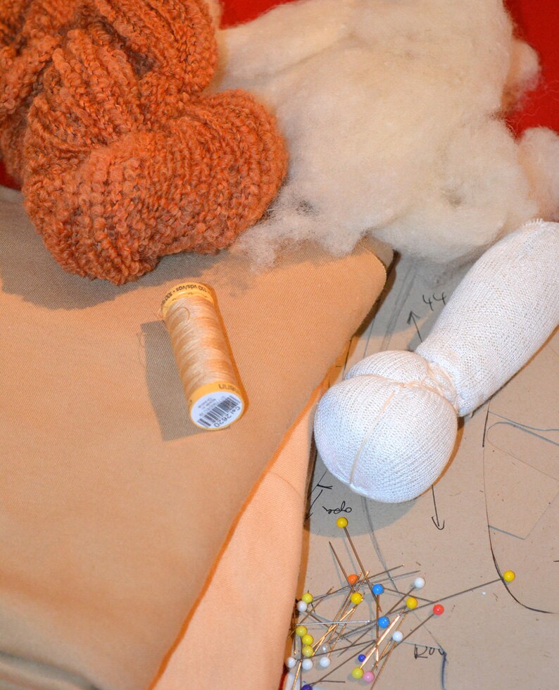 May include: A close-up of a craft project with a spool of thread, a white fabric ball, and a piece of beige fabric. There are also colorful pins and a pattern on the fabric.