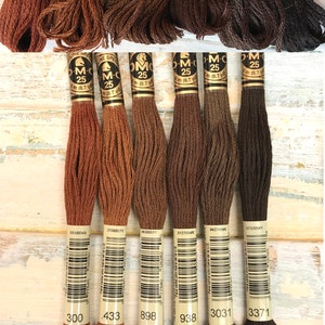 May include: Six spools of DMC embroidery floss in shades of brown, from light to dark. The spools are labeled with DMC numbers: 300, 433, 898, 938, 3031, and 3371.