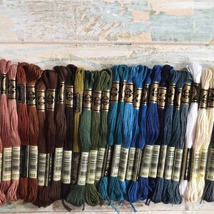 May include: A collection of 25 DMC embroidery floss skeins in various shades of pink, brown, green, blue, gray, white, and black. The skeins are arranged in rows, with the numbers 758, 377, 377, 377, 364, 377, 377, 433, 303, 303, 303, 3071, 520, 936, 330, 378, 803, 803, 803, 176, 743, 443, 317, 379, 31 visible on the labels.