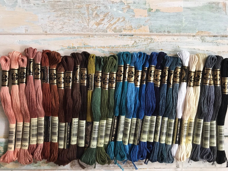 May include: A collection of 25 DMC embroidery floss skeins in various shades of pink, brown, green, blue, gray, white, and black. The skeins are arranged in rows, with the numbers 758, 377, 377, 377, 364, 377, 377, 433, 303, 303, 303, 3071, 520, 936, 330, 378, 803, 803, 803, 176, 743, 443, 317, 379, 31 visible on the labels.