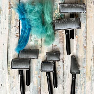 May include: A set of eight metal combs with black handles, used for felting wool. The combs are arranged on a white wooden surface, with a clump of blue and green wool fibres in the top left corner.