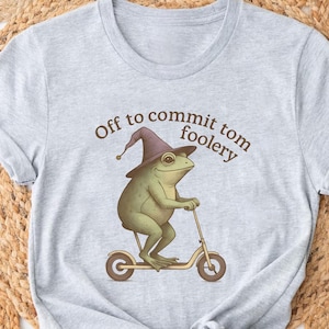 May include: Light gray t-shirt featuring a whimsical illustration of a frog riding a scooter and wearing a wizard hat. The text "Off to commit tomfoolery" is arched above the frog. The shirt is made of soft material.