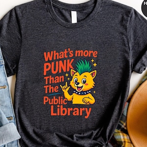 以下が含まれることがあります： 「What's more PUNK Than The Public Library」の赤い文字と、緑のモヒカンヘアの漫画のキツネが描かれた、ヘザーダークグレーのTシャツ。キツネはロックのハンドジェスチャーをしています。