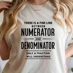 May include: White t-shirt with the text "THERE IS A FINE LINE BETWEEN NUMERATOR AND DENOMINATOR ONLY A FRACTION WILL UNDERSTAND" in black block letters. The shirt is a crew neck style.