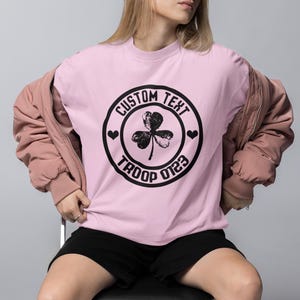 May include: Pink t-shirt with a black circle graphic featuring a shamrock, hearts, and the text "CUSTOM TEXT TROOP 0123".