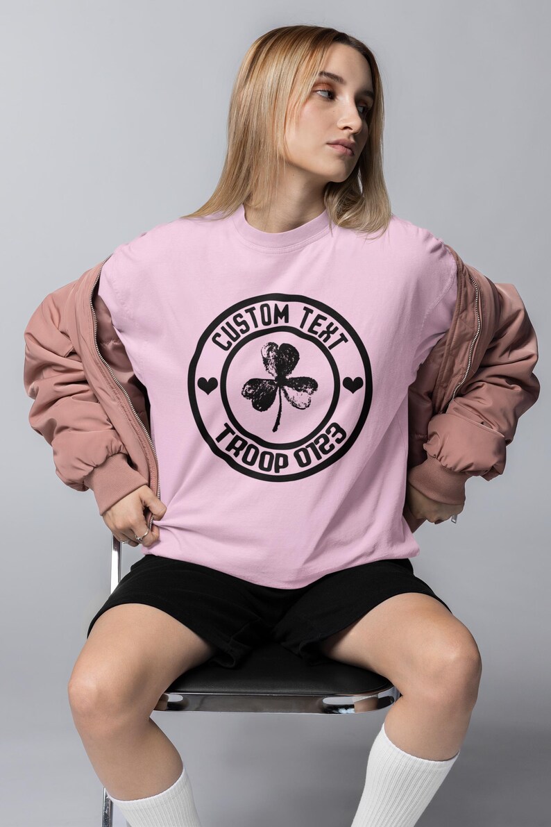 May include: Pink t-shirt with a black circle graphic featuring a shamrock, hearts, and the text "CUSTOM TEXT TROOP 0123".