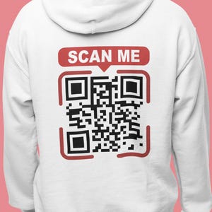 Könnte beinhalten: Weißer Hoodie mit einem roten und schwarzen QR-Code auf der Rückseite. Der QR-Code ist von einem roten Rahmen umgeben und hat den Text "SCAN ME" darüber.