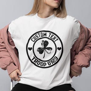 May include: White t-shirt with a black circle design featuring a shamrock, hearts, and the text "CUSTOM TEXT TROOP 0123"