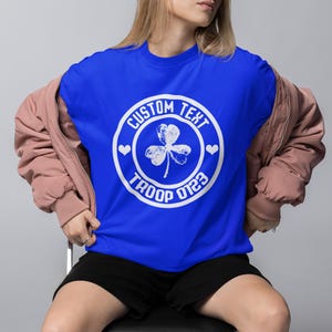 May include: A royal blue t-shirt with a white circle graphic featuring a shamrock, hearts, and the text "CUSTOM TEXT TROOP 0123".
