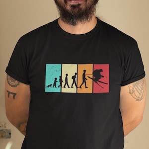 May include: A black t-shirt with a vintage-style graphic of a person skiing. The graphic shows the evolution of skiing, from a baby crawling to a person skiing down a mountain. The graphic is in a rainbow of colors.